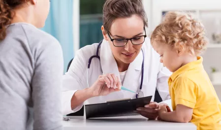 Eine Ärztin erklärt einem jungen Kind etwas. Das Kind hört aufmerksam zu Neben ihnen sitzt eine Frau und schaut zu.