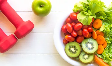 Pinke Hanteln und ein grüner Apfel liegen auf weißem Hintergrund. Eine Schale mit Salat, Tomaten, Kiwi und Karotten steht daneben