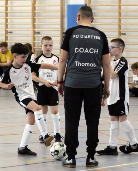 Kinder spielen in einer Halle Fußball. Sie tragen schwarz-weiße Trikots. Im Vordergrund ist außerdem der Trainer zu sehen. Er trägt ein schwarzes Trikot mit der Aufschrift FC Diabetes Coach Thomas