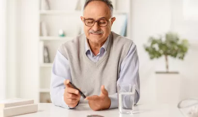 Ein Mann im mittleren Alter misst seinen Blutzucker an seinem Zeigefinger. Er sitzt an einem weißen Tisch, vor ihm steht ein Glas Wasser. Er trägt ein Hemd und einen grauen Pullunder sowie eine dunkle Brille. 