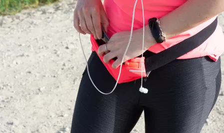Eine Person in schwarzer Sportleggins und pinkem Sportoberteil steckt ein Handy in eine Bauchtasche. Sie trägt eine Smartwatch und Kopfhörer.